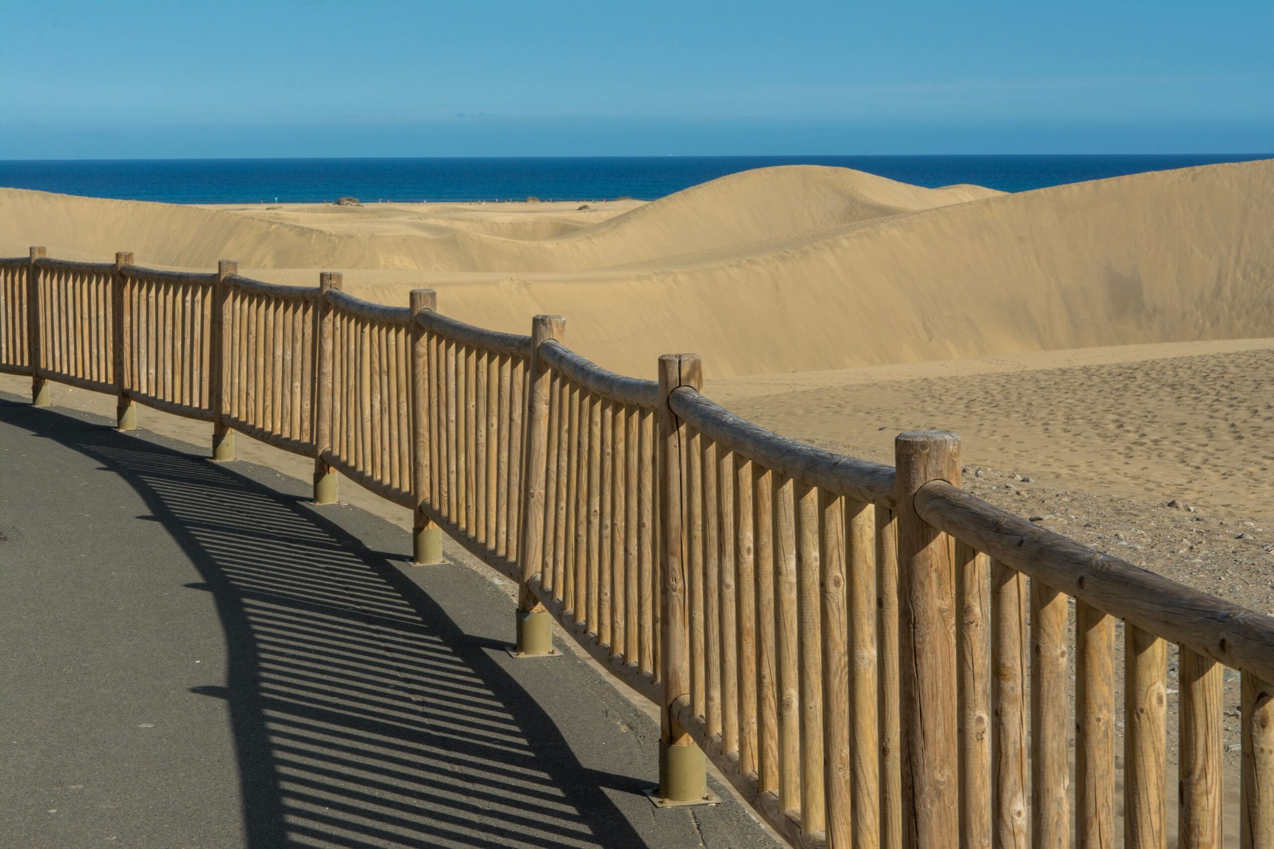 A wooden walkway with shadows leads to the sandy dunes and blue ocean in Maspalomas, Spain.